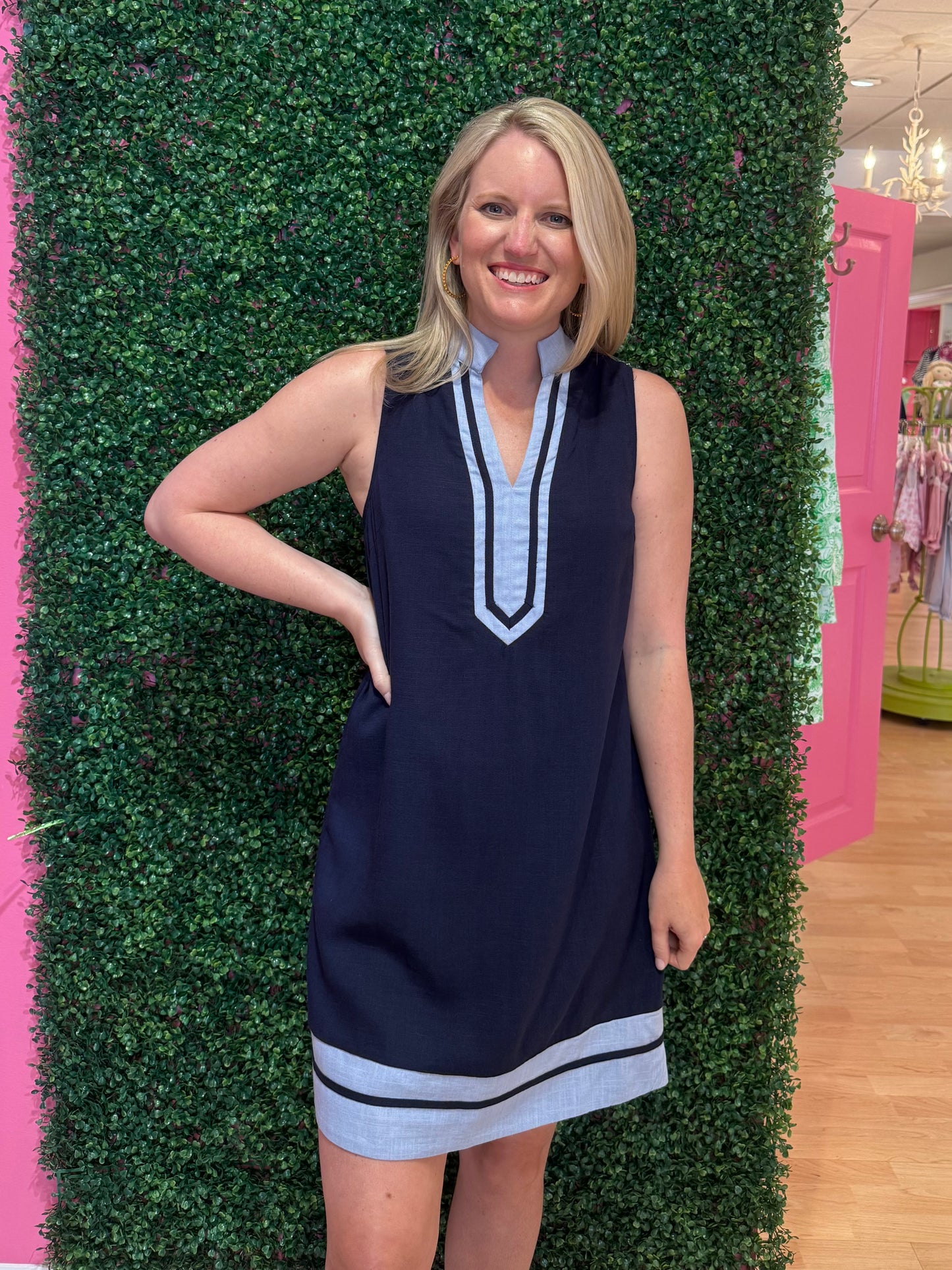 Woman in a navy dress with light blue trim standing against a green bush wall