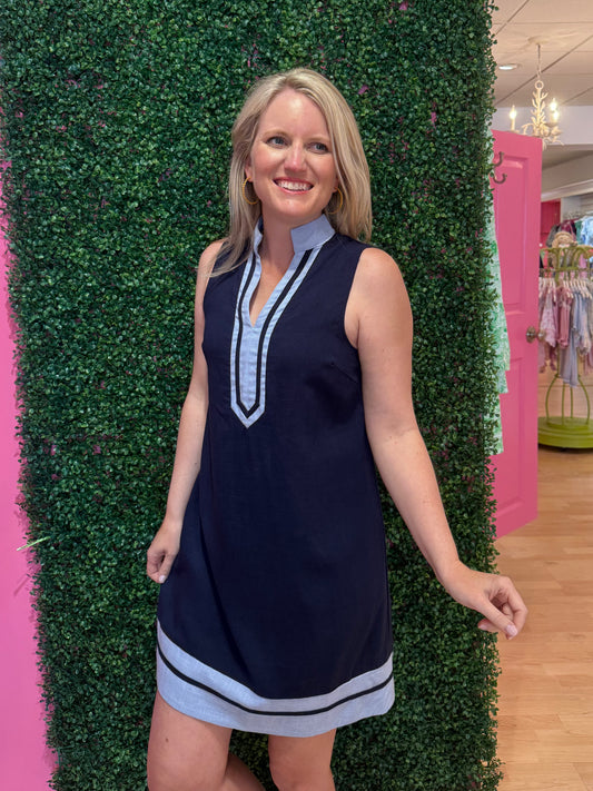 Woman wearing a navy dress with light blue trim standing in front of a green hedge.